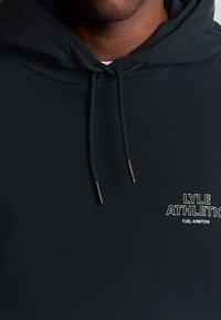 Felpa verde scuro con cappuccio, dotata di tasca a marsupio frontale, lacci regolabili e logo testuale bianco: "LYLE ATHLETIC FUEL AMBITION." Texture liscia.