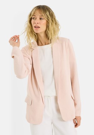Femme blonde en blazer rose clair, top blanc et pantalon blanc, tenant une mèche de cheveux et regardant vers sa gauche.