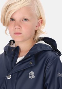 Tamsiai mėlyna nepralaidi vandeniui striukė su aukštu apykakle, sidabriniais elementais ir logotipo detale. Yra užtrauktukų uždarymas ir vidinė pilka pamušalas.