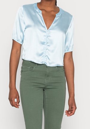 Person trägt eine hellblaue Satinbluse mit kurzen Ärmeln und Knopfdetails sowie eine hochtaillierte olivgrüne Hose.