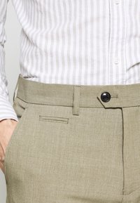 Licht khaki broek met een gladde textuur, voorzien van een knoopsluiting, riemlussen en een verborgen zakdetails op de tailleband.