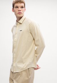 Beige blouse met lange mouwen, voorzien van een geometrisch patroon en een subtiele glans, met een puntige kraag en een zwart "BOSS" logo op de borst.