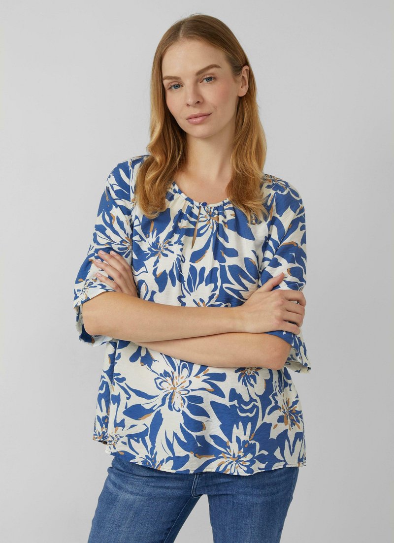 Frau mit hellbraunen Haaren, die eine blau-weiße Bluse mit Blumenmuster und blaue Jeans trägt, steht mit verschränkten Armen vor einem schlichten Hintergrund.