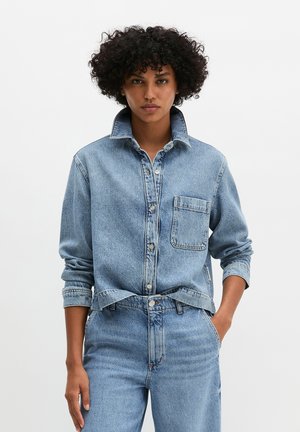 Personne aux cheveux bouclés portant une chemise en jean bleu clair avec le col relevé, rentrée dans un jean taille haute assorti, debout avec une main dans la poche.