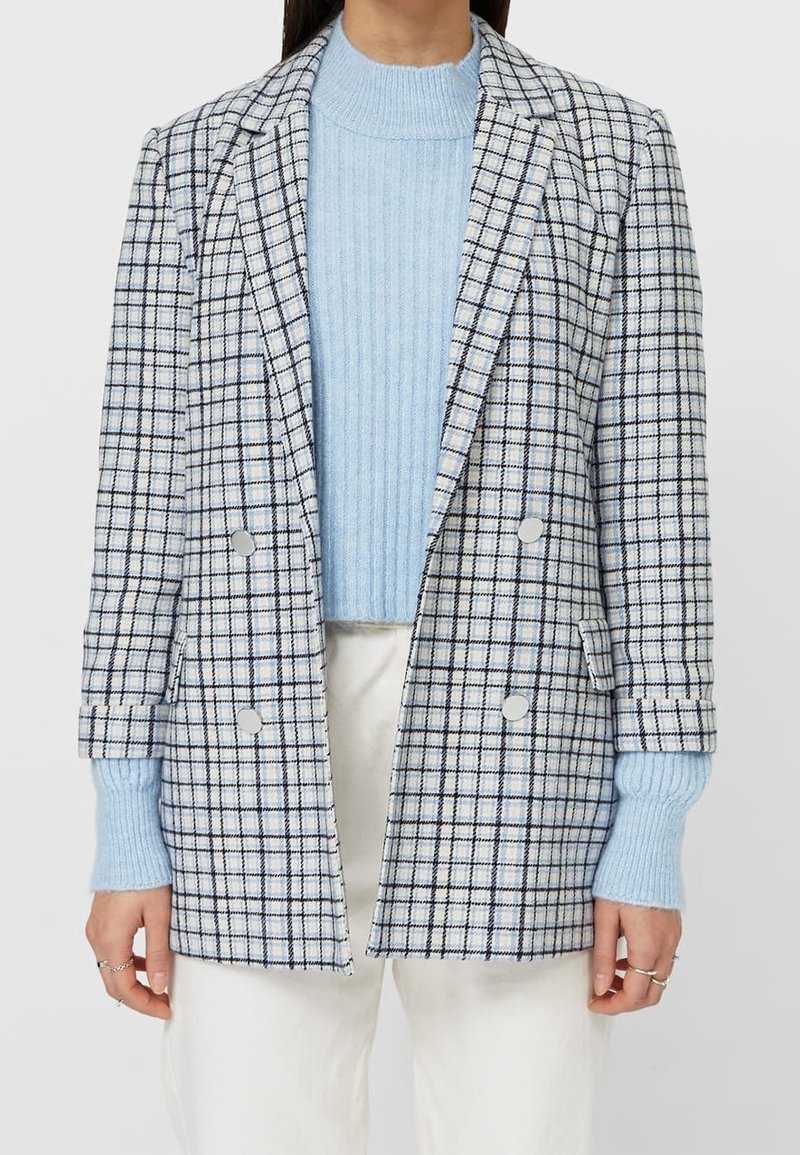 Femme portant un pull côtelé bleu clair sous un blazer croisé à carreaux blanc et marine avec un pantalon blanc, mains le long du corps.