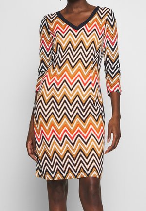Femme portant une robe mi-longue à manches trois-quarts avec un motif zigzag multicolore en noir, orange, rouge et blanc.