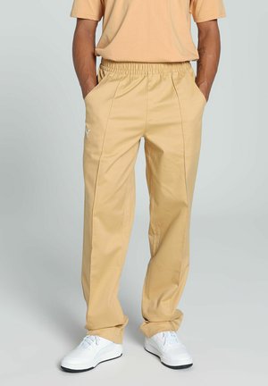 Beige relaxed-fit pants with an elastic waistband and side pockets. Features a subtle brand logo in white on the left thigh.