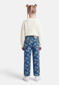 Παιδί με δύο πλεξούδες, φοράει λευκό πλεκτό πουλόβερ και μπλε τζιν με ανθικό σχέδιο, βλέποντας προς τα πίσω σε απλό λευκό φόντο.