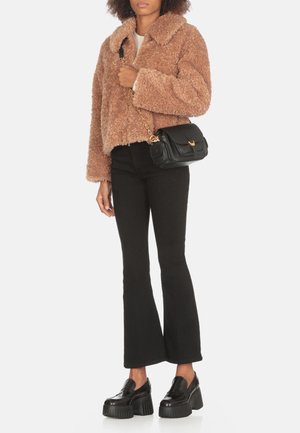 Giacca corta in finta pelliccia marrone con colletto, jeans neri a zampa, e loafers neri con suola spessa. Piccola borsa a tracolla nera con dettagli dorati.