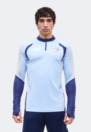 Muškarac u svijetlo plavoj i tamnoplavoj sportskoj majici dugih rukava s četvrtastim patentnim zatvaračem i Adidas logom, stoji nasuprot jednobojnoj svijetloj pozadini.
