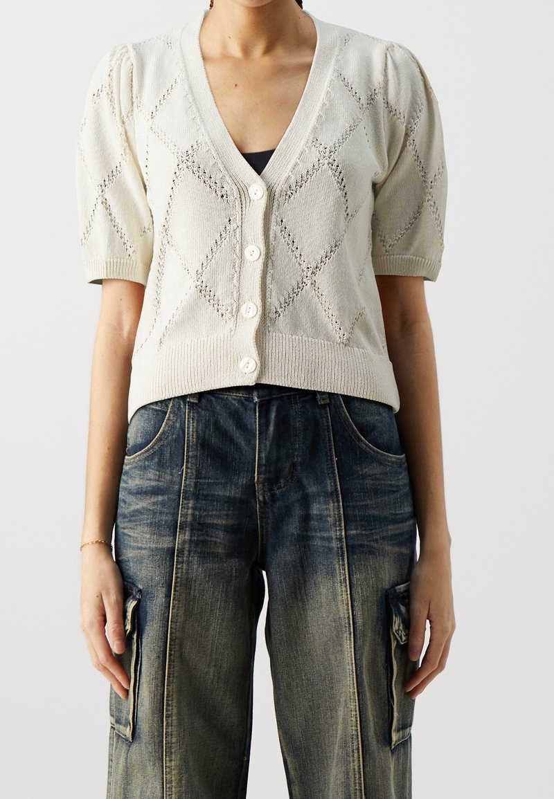 Femme portant un cardigan en maille crème à manches courtes avec des motifs en losange et un jean cargo large délavé.