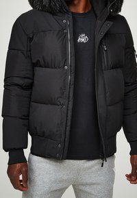 Czarna pikowana zimowa kurtka z kapturem wyłożonym futerkiem, zapinana na zamek, z elastycznymi mankietami i bocznymi kieszeniami. Noszona na czarnej koszulce i szarych spodniach dresowych.