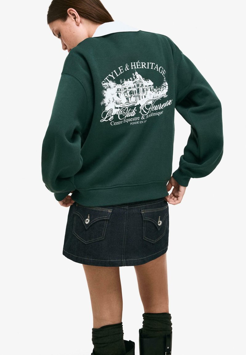 Zelena jopica s grafiko "STYLE & HÉRITAGE" na hrbtni strani, v kombinaciji s temno denim mini krilom, ki ima dva zadnja žepa in podrobnosti v stiku.