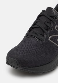 Primer plano de una zapatilla de running negra de malla transpirable con cordones, puntera reforzada y suela texturizada sobre un fondo blanco.