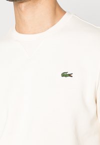 Primer plano de una persona que lleva un sudadera blanca con un pequeño logo de cocodrilo verde bordado en el pecho.
