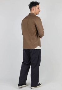Chemise marron à boutons, avec une coupe ajustée et des fentes sur les côtés, assortie à un pantalon noir ample et des baskets noires. Texture lisse sur l'ensemble.