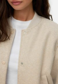 Giacca bomber beige con una trama morbida, colletto a coste, chiusure con bottoni a pressione e un design semplice, indossata sopra una camicia bianca.