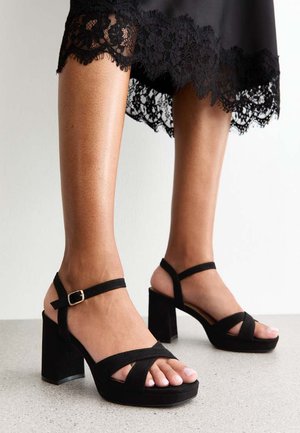 Jambes portant des sandales noires à talons hauts avec des brides à la cheville et une robe noire avec un bord en dentelle à l'ourlet.