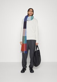 Pull en gris clair, coupe oversize. Écharpe en laine colorée et à motifs, avec des franges. Pantalon ample gris foncé. Sac noir avec poignées. Chaussures noires.