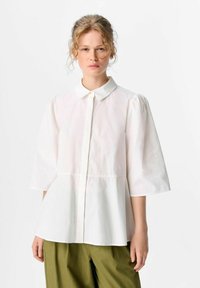 Weiße Bluse mit Kragen und kurzen Puffärmeln, hat eine Naht unter der Brust und eine lässige Silhouette. Der Stoff wirkt glatt und leicht.