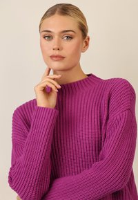 Maglione lavorato a maglia di un vivace colore viola, caratterizzato da una trama a coste, colletto alto e maniche ampie per una vestibilità rilassata.
