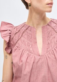 Γυναίκα με ροζ μπλούζα με υφή και βολάν στα μανίκια και χρυσό σκουλαρίκι στεφάνι, που δείχνει μέρος τατουαρισμένου χεριού.