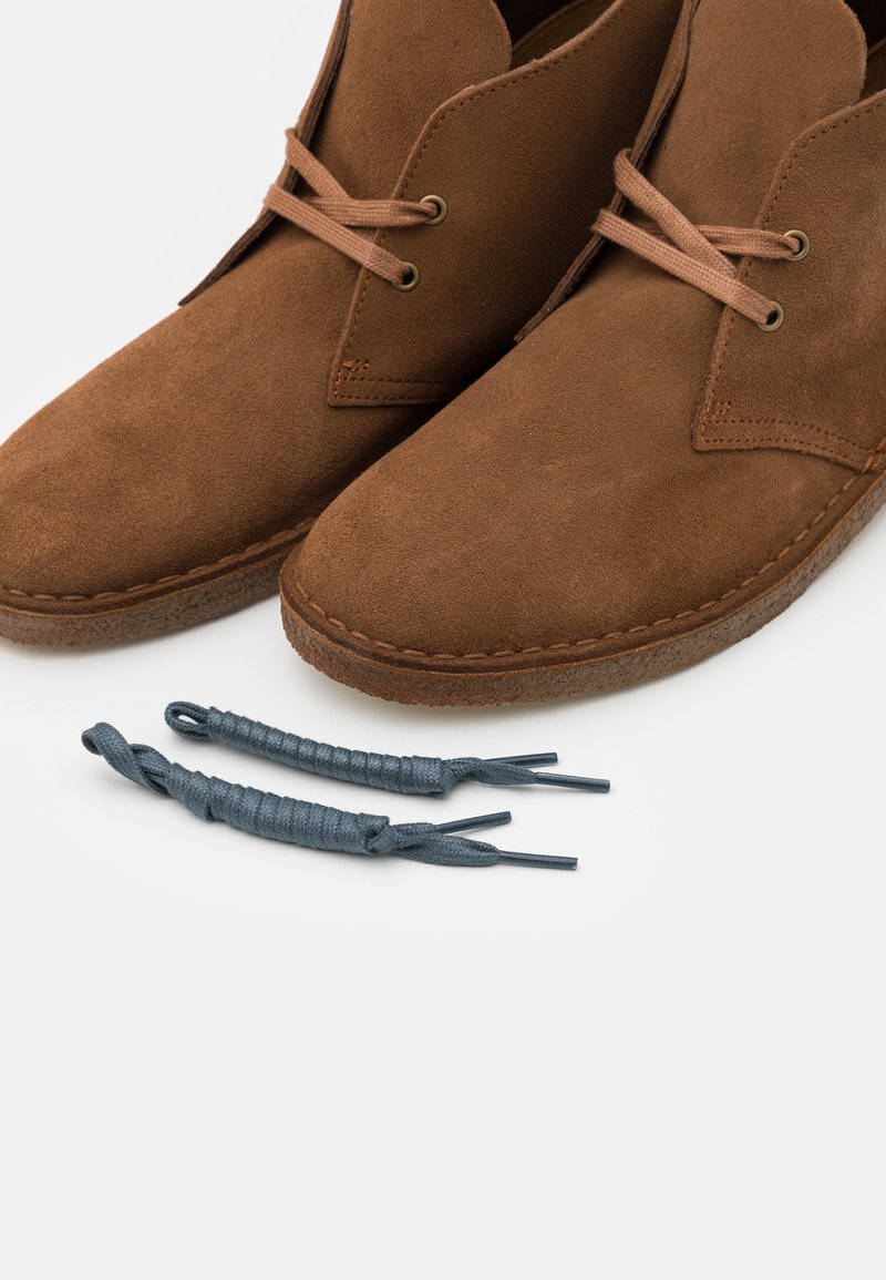 Zapatos de ante marrón con punta redondeada y detalles de costura, acompañados de dos cordones texturizados azul oscuro.