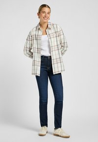 Lichte geruite button-up shirt in beige, blauw en grijs, gedragen over een witte top; gecombineerd met donkere skinny jeans en lichte sneakers.