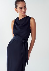 Mornariće plava haljina bez rukava s izrezom u obliku cowl, detaljem remena oko struka i glatkom teksturom tkanine. Elegantna i oblikovana silueta.