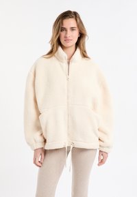 Kremowa kurtka polarowa z wysokim kołnierzem, zamkiem błyskawicznym i dużymi przednimi kieszeniami. Charakteryzuje się luźnym krojem i ściągaczem u dołu.