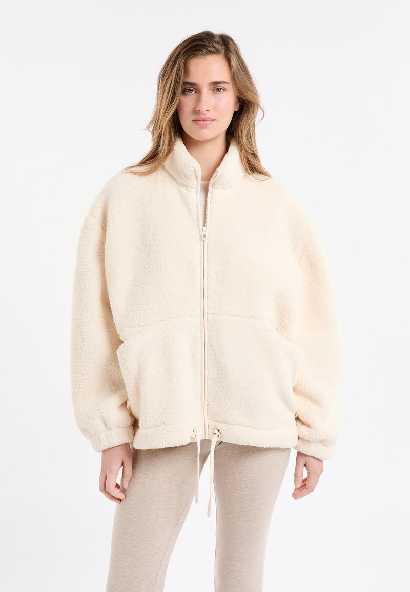 Kremowa kurtka polarowa z wysokim kołnierzem, zamkiem błyskawicznym i dużymi przednimi kieszeniami. Charakteryzuje się luźnym krojem i ściągaczem u dołu.