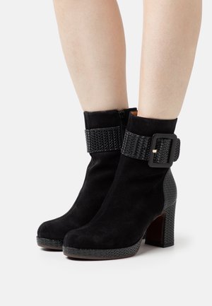 Bottes cheville en daim noir avec une sangle tissée texturée et une boucle rectangulaire, dotées d'un talon bloc et d'un design à bout arrondi.