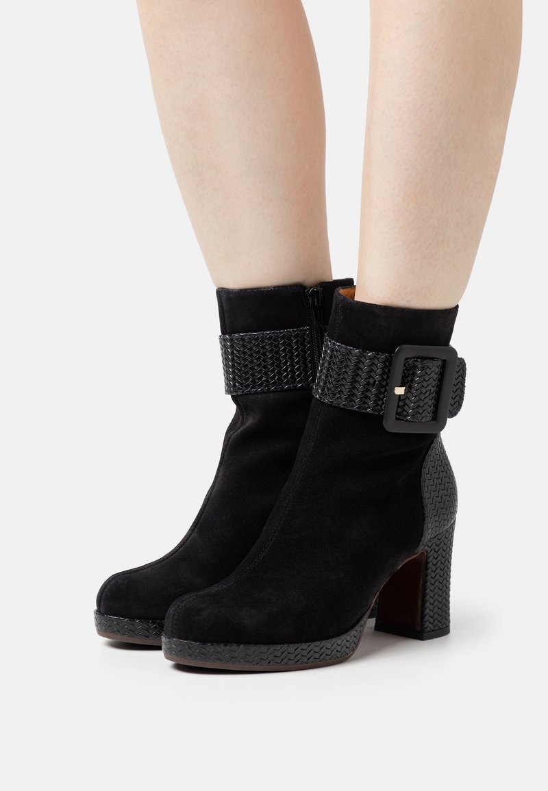 Bottes cheville en daim noir avec une sangle tissée texturée et une boucle rectangulaire, dotées d'un talon bloc et d'un design à bout arrondi.