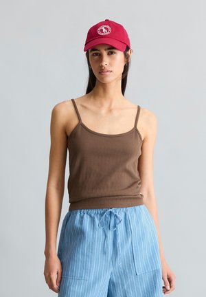 Jeune femme portant une casquette de baseball rouge, un débardeur marron et un pantalon bleu rayé à cordon sur un fond gris uni.