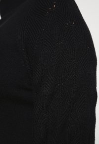 Gestrickter schwarzer Pullover mit einem strukturierten Design, das wellenförmige Muster an den Ärmeln zeigt. Das Material wirkt weich und warm.