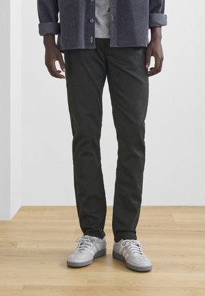 Dunkelgrüne, schlank geschnittene Hose aus Baumwolle, kombiniert mit einem grauen Shirt und weißen Sneakern. Holzboden und neutrale Wand im Hintergrund.