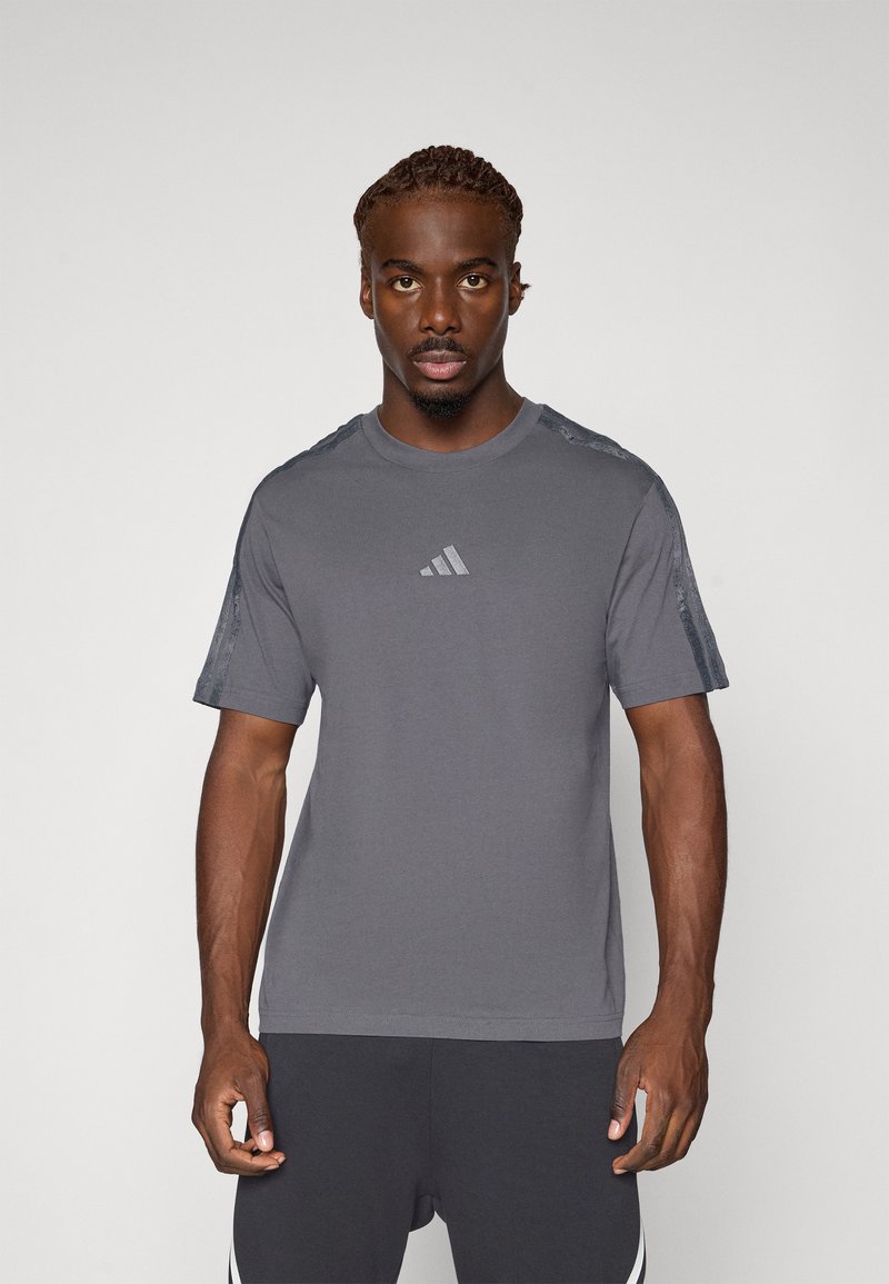 T-shirt gris à manches courtes avec un col rond, présentant un motif texturé sur les épaules et un logo discret sur la poitrine.