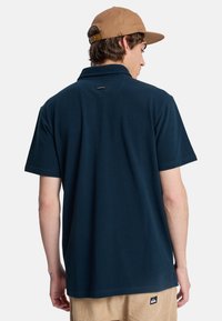 Mørkeblå polo-shirt med en blød tekstur, korte ærmer og krave. Har et lille logo-patch bagpå og er parret med beige bukser.