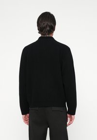 Moški s temnimi lasmi, oblečen v črno rebrasto pulover in črne hlače, stoji z zavrtanim hrbtom proti kameri na belem ozadju.