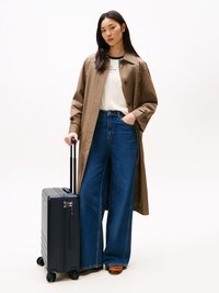 Valigia rigida blu navy con manico estraibile e quattro ruote, abbinata a un cappotto trench beige e jeans in denim a gamba larga.