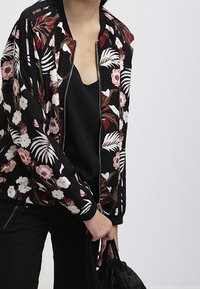 Person iført en sort blomstret zip-up jakke over en sort top, der holder en sort snørerpose mod en ensfarvet grå baggrund.