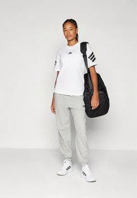 Balta īsroku T-krekls ar melnu logotipu un svītrām, pelēkas sporta bikses un baltas sporta kurpes, nēsājot melnu sporta somu.