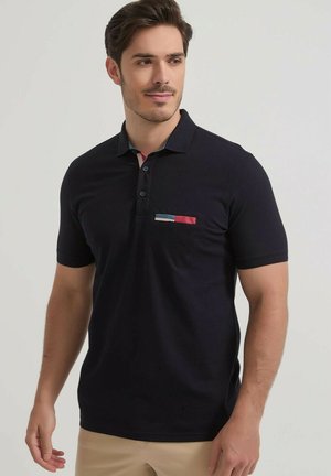 Homme portant un polo noir à manches courtes avec des détails rouges, bleus et blancs sur le col et la poche poitrine.