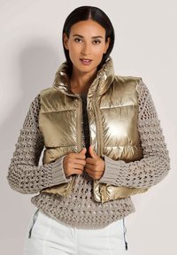 Metallisch goldene Pufferweste mit hohem Kragen. Getragen über einem hellgrauen, offen gestrickten Pullover. Reißverschluss und abgerundeter Saum. 