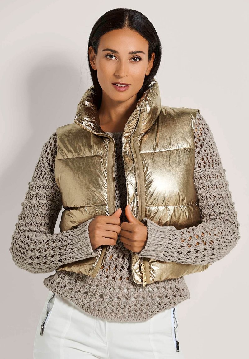 Metallisch goldene Pufferweste mit hohem Kragen. Getragen über einem hellgrauen, offen gestrickten Pullover. Reißverschluss und abgerundeter Saum.