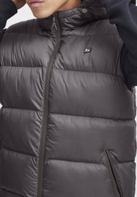 Sivi softshell prsluk s visokim ovratnikom, zatvaranjem na zip i elastičnom vezicom. Ima dva bočna džepa i mali logo na prsima.