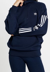 Sweatshirt zippé bleu marine avec col montant, logo Adidas blanc sur la poitrine et trois bandes blanches le long des manches. Texture douce.