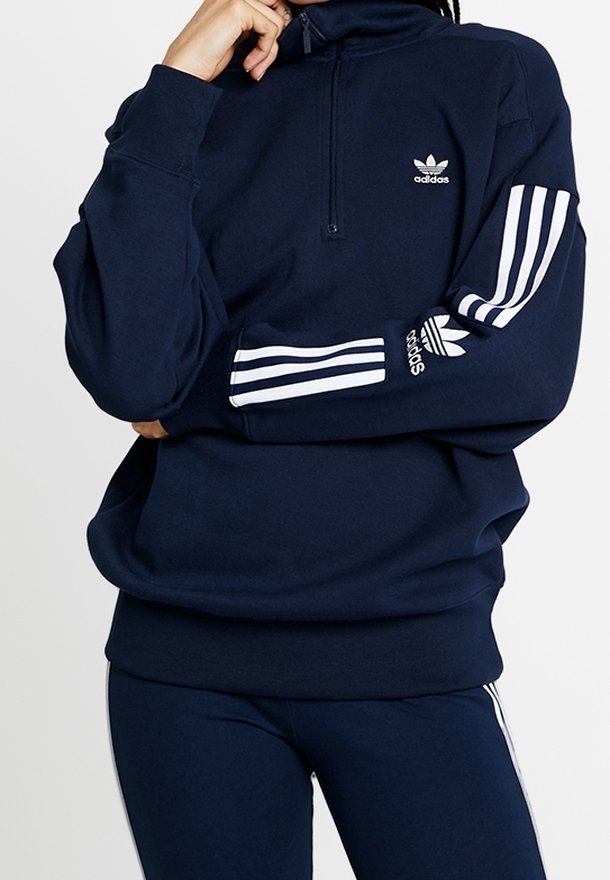 Sweatshirt zippé bleu marine avec col montant, logo Adidas blanc sur la poitrine et trois bandes blanches le long des manches. Texture douce.