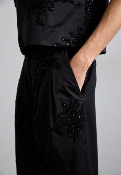 Juodas komplektas, sudarytas iš trumpų palaidinių ir kelnių, su sudėtingais juodais blizgučiais dekoruotais gėlių raštais ir glotnia satino tekstūra.