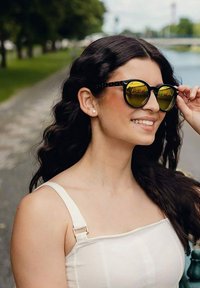 Černé sluneční brýle se žlutými skly, s geometrickým vzorem. Modelka má vlnité vlasy a nosí světlý top se špagetovými ramínky.